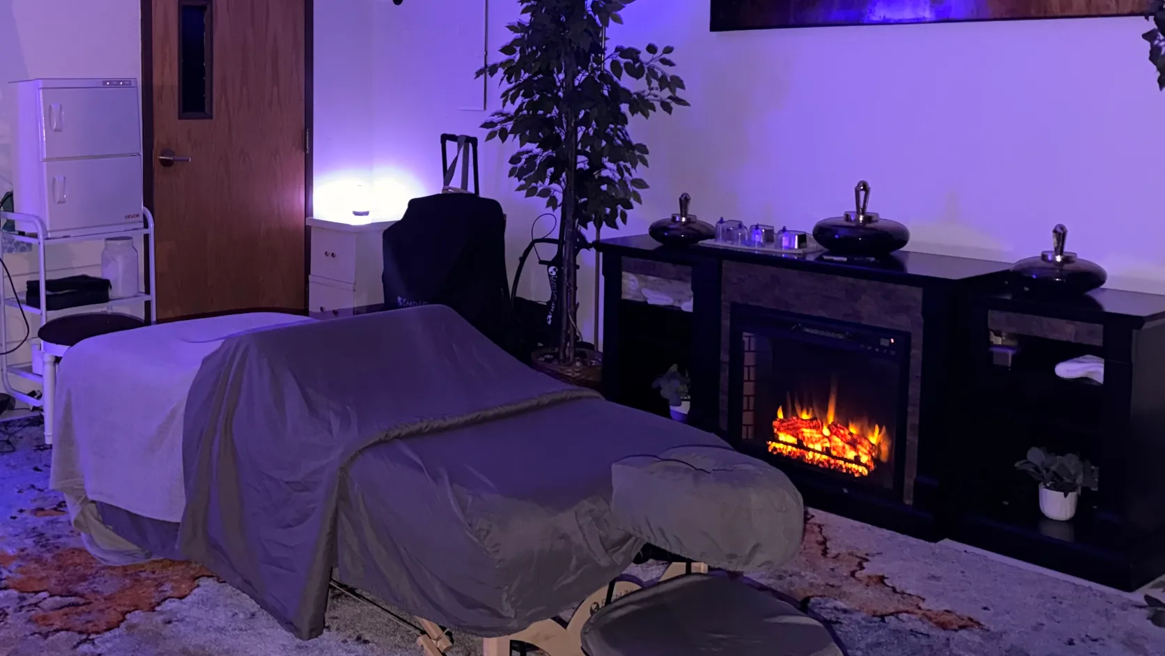 The current M.M.M. Serenity massage room with a treatment table, fireplace, and purple ambient lighting